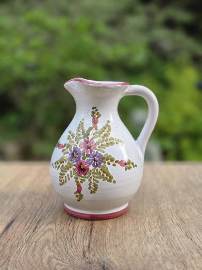 Kleiner Vintage-Krug aus glasierter Keramik mit bauchiger Silhouette, zartem rosa Rand und handgemaltem Blumendekor in Rosa, Lila und Grün. Die kompakte Form mit schmalem Hals und feinem Ausguss wirkt ruhig und dekorativ. Stilistisch Mid Century, vermutlich aus dem süddeutsch-österreichisch-italienischen Raum, datierbar in die 1950er bis 1970er Jahre. Guter Zustand mit herstellungsbedingten Lasurfehlern, ohne Beschädigungen.