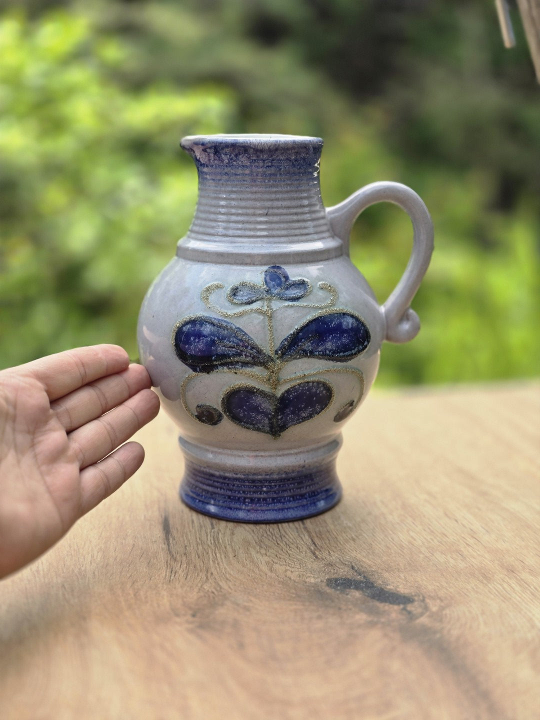 Strehla Keramikkrug neben einer Hand zur Größenansicht, mit floralem Dekor, blauem Fuß und heller Glasur.