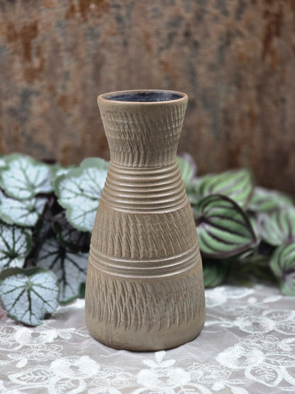 Keramikvase mit schlanker Form, braungrauer Oberfläche, Kerbschnittdekor und Rillenbändern auf weißer Spitze vor Pflanzen