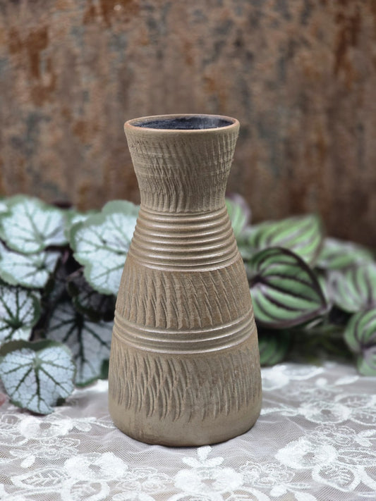 Keramikvase mit schlanker Form, braungrauer Oberfläche, Kerbschnittdekor und Rillenbändern auf weißer Spitze vor Pflanzen