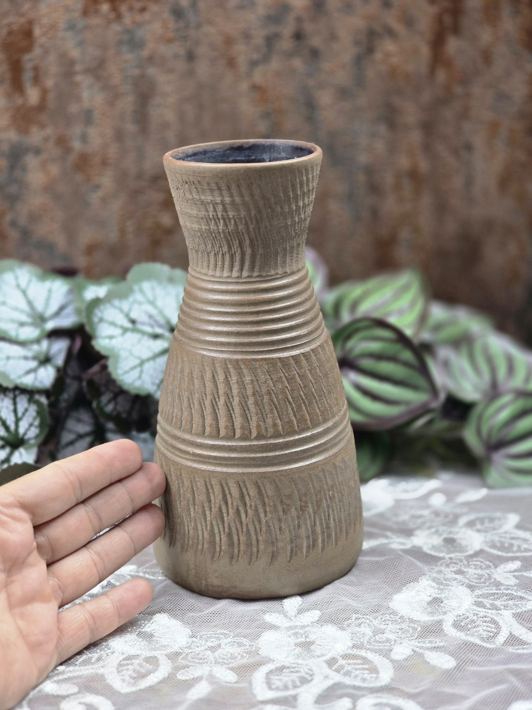 Keramikvase mit schmaler Taille, eingeritztem Muster, umlaufenden Rillen und matter braungrauer Glasur zwischen Blattpflanzen