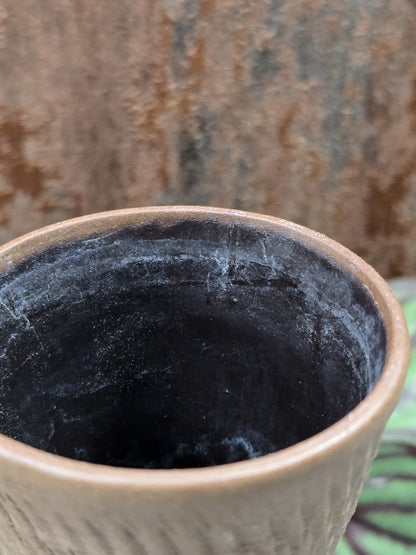 Keramikvase in Frontalansicht mit breitem Stand, engem Hals, Kerbschnittdekor und braungrauer Oberfläche vor Pflanzen
