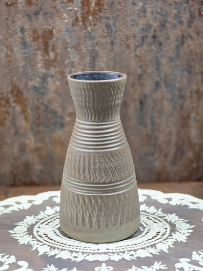Keramikvase mit rosa Kunstblumen, braungrauer Oberfläche, Rillenbändern und Kerbschnittdekor zwischen Blattpflanzen