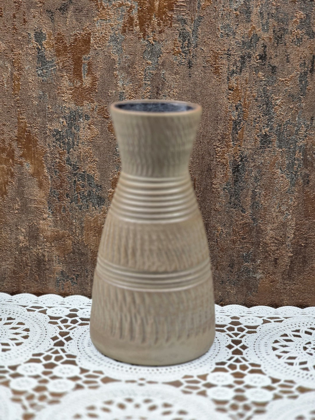 Keramikvase mit schlanker Form, Rillenbändern und Kerbschnittmuster auf weißer runder Spitzendecke vor Strukturwand