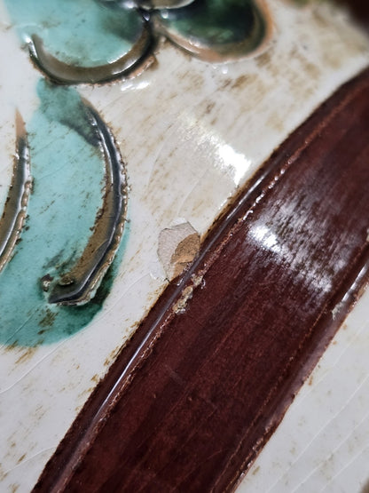 Keramikvase mit floralem Reliefdekor und sichtbarem Chip neben braunem Zierband in Detailaufnahme