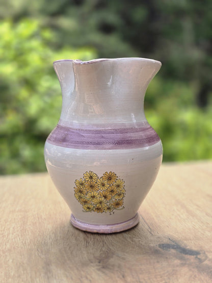 Handgemachter Keramikkrug mit gelbem Blumendekor, violettem Band und heller Glasur vor grünem Hintergrund.