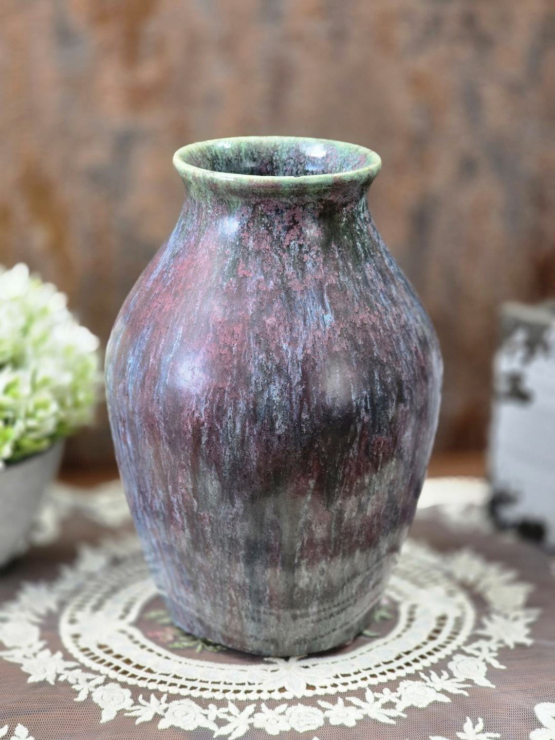 Vintage Keramikvase mit mehrfarbiger Laufglasur in Grün, Mauve und Blau dekorativ auf einer weißen Spitzendecke arrangiert.