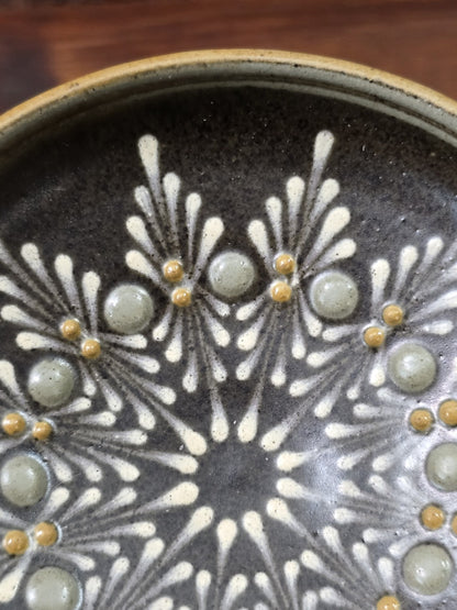 Keramikteller mit dunkelbrauner Glasur, weißen Strahlenmotiven und erhabenen grauen sowie gelben Punkten in Detailaufnahme