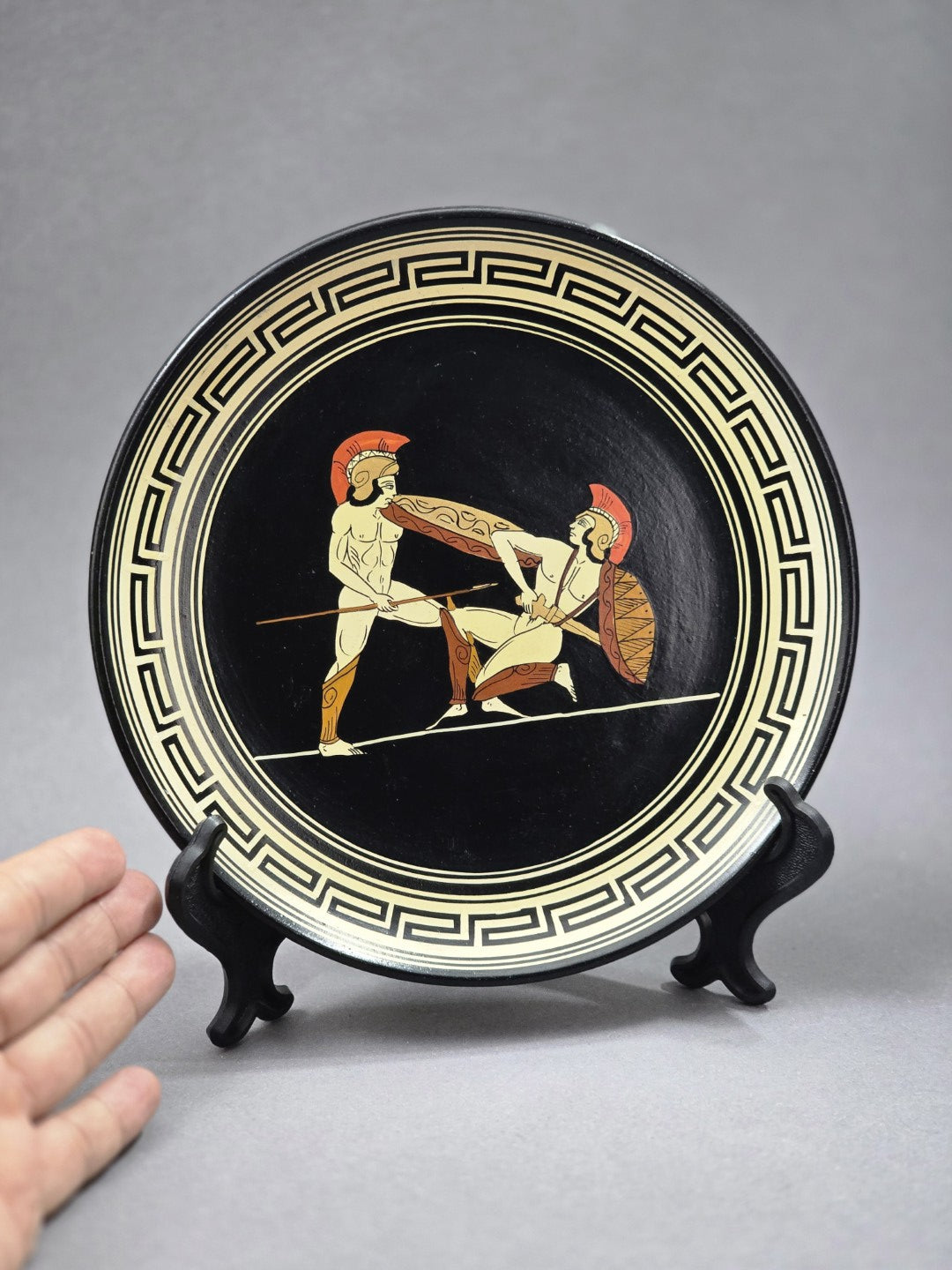 Vintage Wandteller mit zwei antiken Kriegerfiguren, schwarzem Grund und cremefarbenem Mäanderrand, mit Hand als Größenvergleich