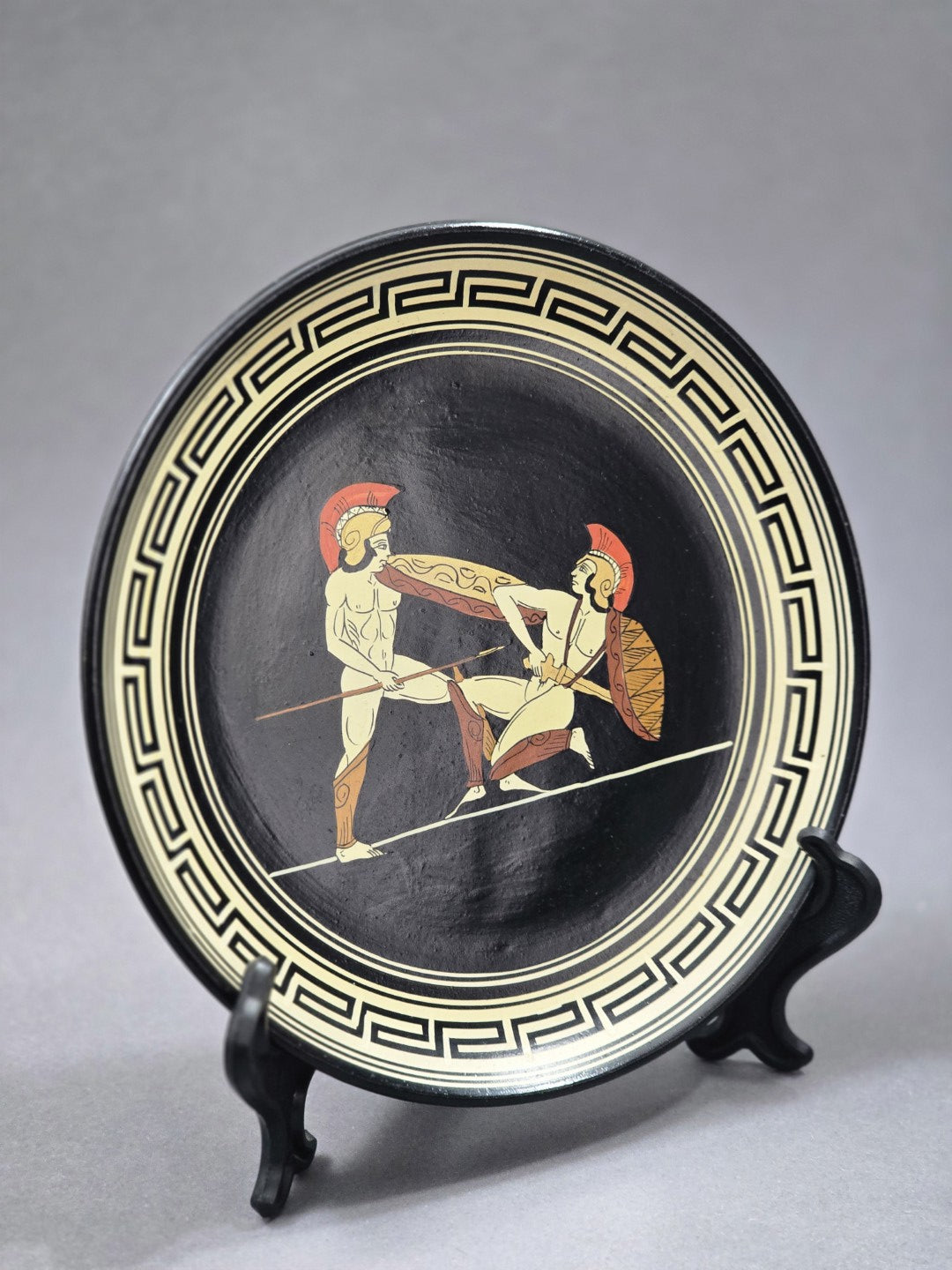 Vintage Wandteller mit griechischer Kampfszene in Creme, Rot und Ocker auf schwarzem Grund, schräg von links fotografiert