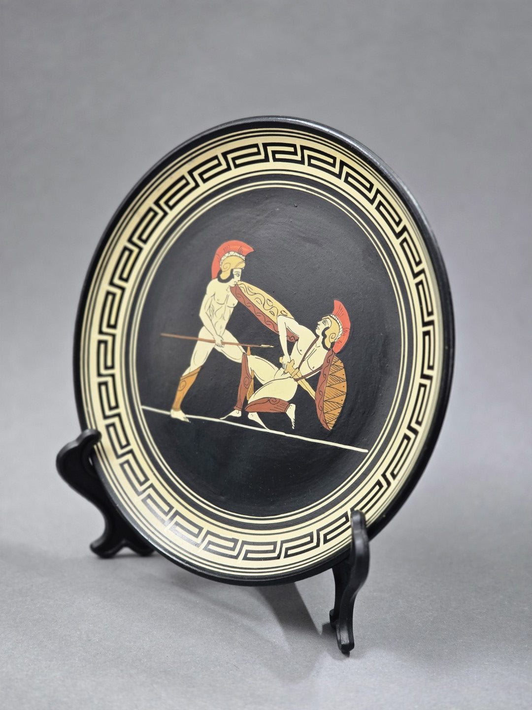 Vintage Wandteller mit zwei Kriegern und geometrischem Mäanderdekor, seitlich schräg auf schwarzem Tellerständer gezeigt