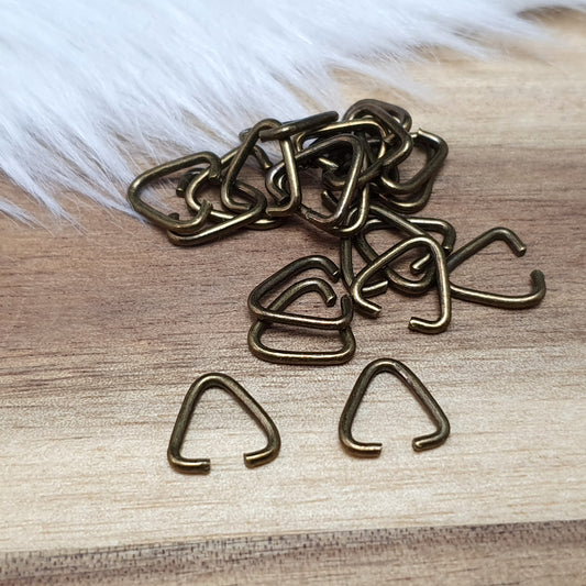 Das Bild zeigt ein Set mit 40 offenen Triangel-Ösen aus antik bronzefarbener Eisenlegierung, die eine Höhe von 9 mm haben. Die Ösen sind in Form eines gleichseitigen Dreiecks gestaltet, wobei eine Seite offen ist, um das Einfügen von Schmuckkomponenten zu erleichtern. Die matte, antik bronzefarbene Oberfläche verleiht den Ösen einen charmanten Vintage-Look, der ideal für verschiedene Schmuckherstellungsprojekte ist. Das Set ist übersichtlich angeordnet und hebt die Vielseitigkeit dieser Ösen hervor, die per